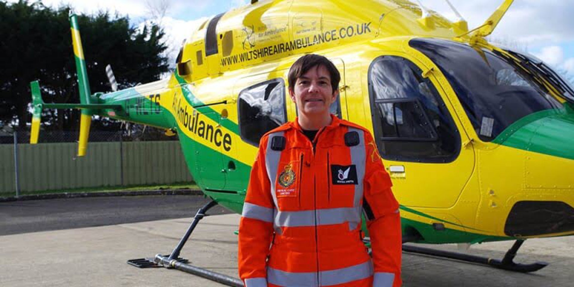 A critical care doctor wearing a flight suit in front of the Wiltshire Air Ambulance helicopter. 