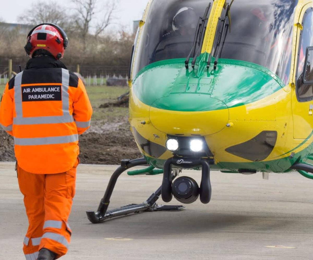 A paramedic wearing an orange flight suit, coat and red flight helmet walking towards the nose of the helicopter.