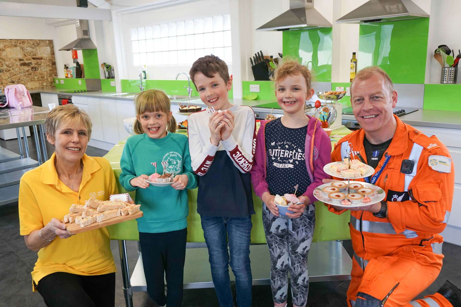 A group photo taken in a professional kitchen with children holding cakes alongside staff and critical care paramedic.