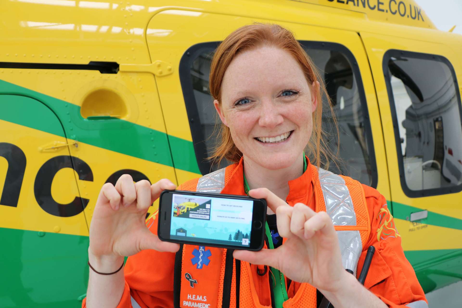 A critical care paramedic stood holding a phone with the app Helifun on screen.