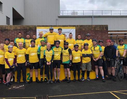 A group of cyclists stood outside a factory ready to take part in a cycle ride. They are wearing yellow t-shirts and cycling helmets.