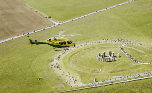 The helicopter flying over Stone Henge with lots of visitors walking around.