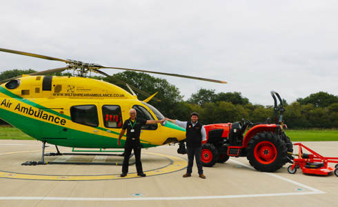An orange Kubota tractor and trailer parked on the helipad next to the helicopter. There is a staff member from WAA and Howard & Sons handing over the keys.