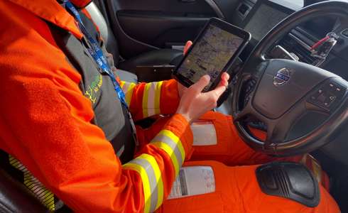 A paramedic wearing an orange flight suit sat in the drivers seat of the critical care car. They are holding an iPad which shows an image of a map.