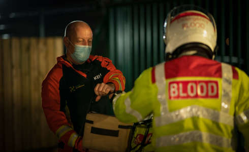 A paramedic who is being handed a delivery box of blood products from a biker wearing a high vis jacket and a helmet.