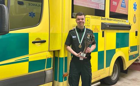 Leon in uniform holding the World Irish Dance Champion trophy, with a South Western Ambulance Service NHS Foundation Trust vehicle