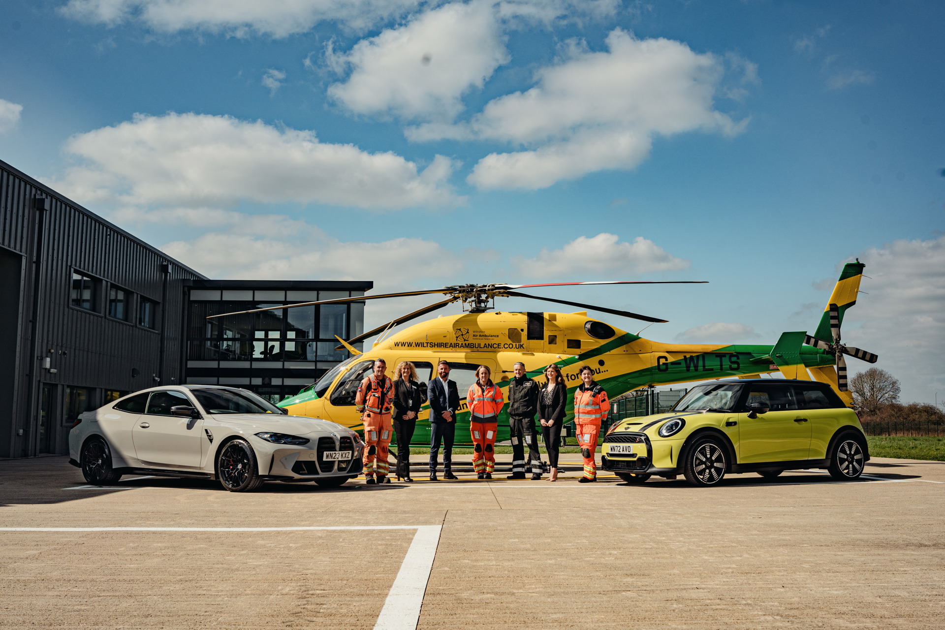 The Wiltshire Air Ambulance helicopter on the helipad with a yellow mini and grey BMW parked alongside