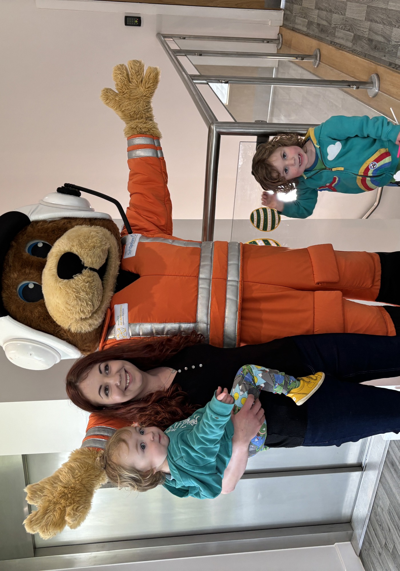 An orange paramedic bear mascot posting with an adult, holding a small child, and another child standing with them