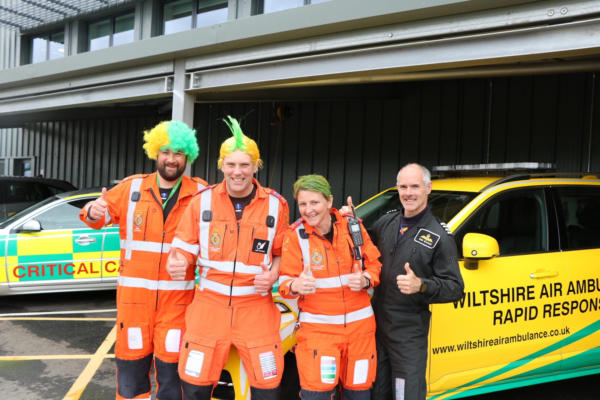 3 paramedics wearing orange flight suits, wearing yellow and green wigs, and a pilot wearing a black flight suit with their thumbs up