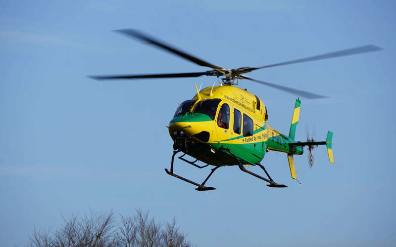 The yellow and green helicopter in flight against a blue sky with trees.