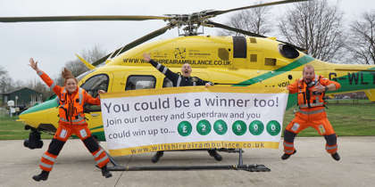 Two paramedics and a pilot jumping and smiling in front of the helicopter on a helipad. They are holding a large white and yellow banner advertising the Lottery Superdraw.