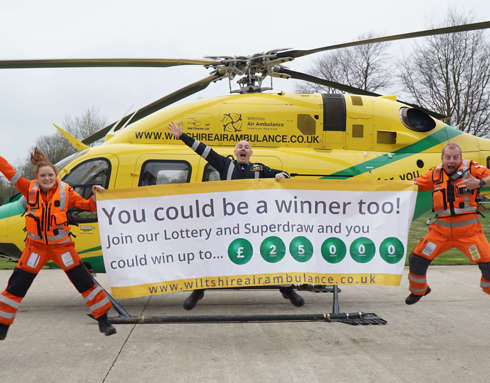 Two paramedics and a pilot jumping and smiling in front of the helicopter on a helipad. They are holding a large white and yellow banner advertising the Lottery Superdraw.