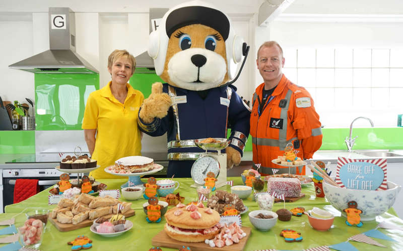 Paramedic posing with mascot Marsha and charity team member, with a table full of cake