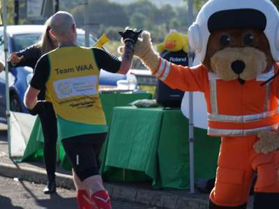 A runner wearing a Wiltshire Air Ambulance branded yellow and green running vest, highfiving our paramedic mascot Wilber, whilst WAA's volunteers cheer them on