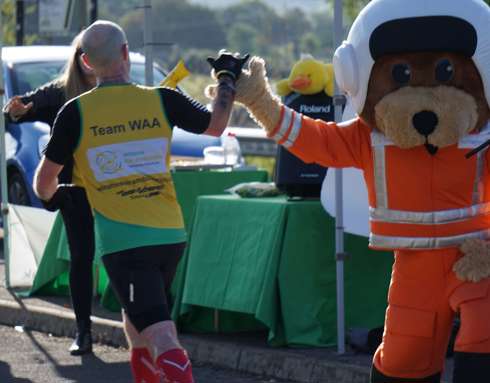 A runner wearing a Wiltshire Air Ambulance branded yellow and green running vest, highfiving our paramedic mascot Wilber, whilst WAA's volunteers cheer them on