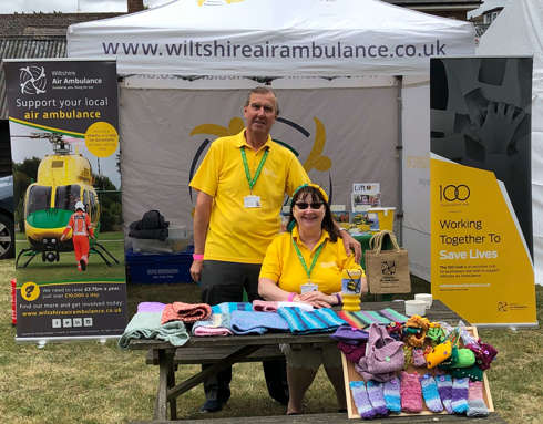 A pair of volunteers wearing yellow polo shirts. They are in front of the charity's gazebo with a range of knitted merchandise placed on the table in front of them.