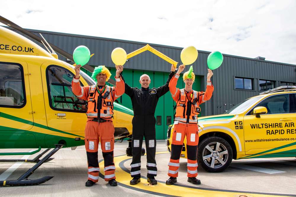 Three members of Wiltshire Air Ambulance aircrew wearing yellow and green wigs and holding balloons in front of the Bell-429 helicopter and critical care car.