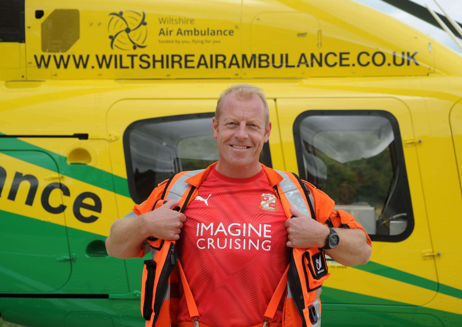 Critical care paramedic wearing a flight suit and Swindon Town Football shirt underneath stood in front of the Bell-429 helicopter.