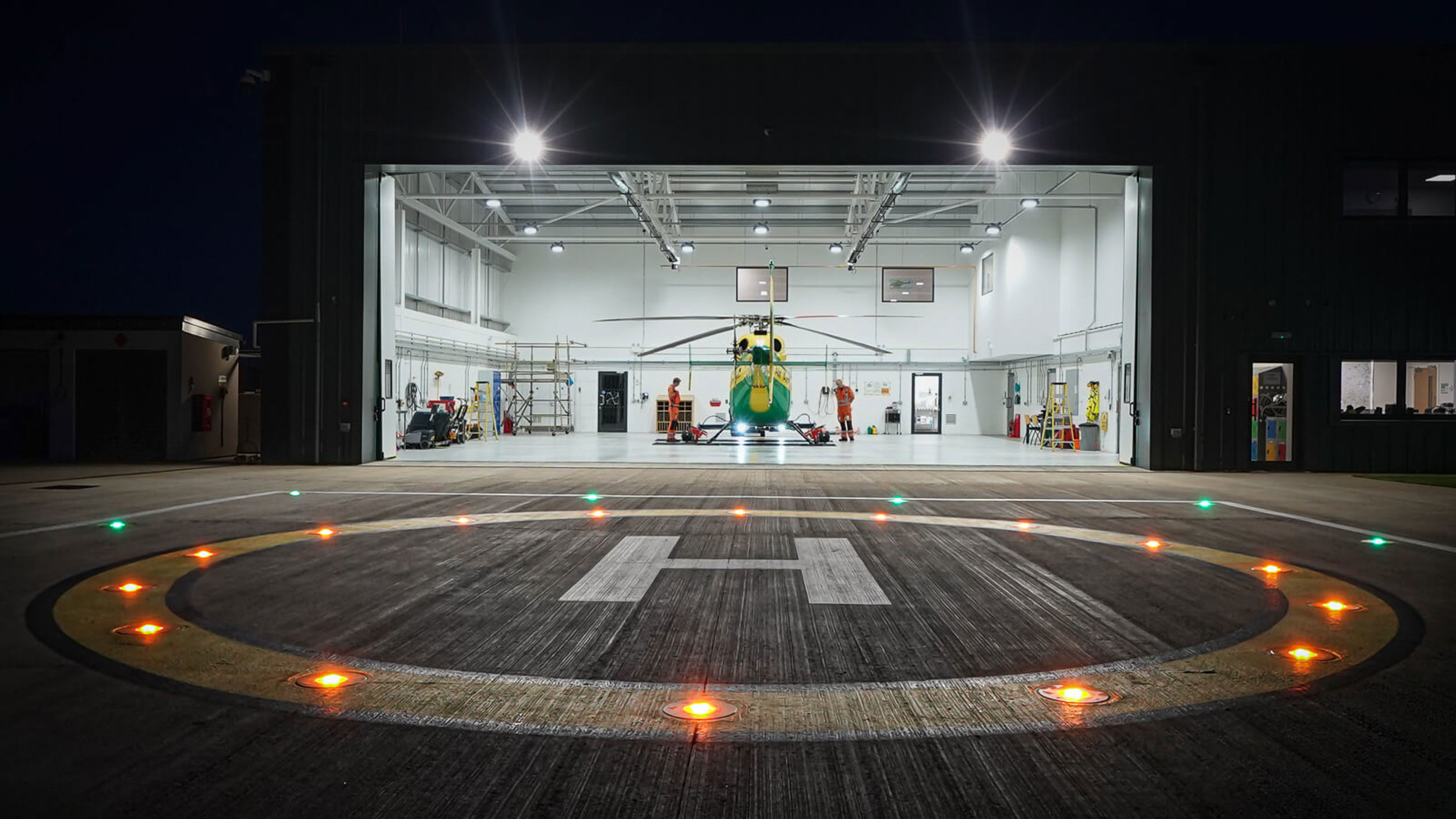 The Wiltshire Air Ambulance helipad at night with yellow and green lights shining against the bright white lights inside the hangar.