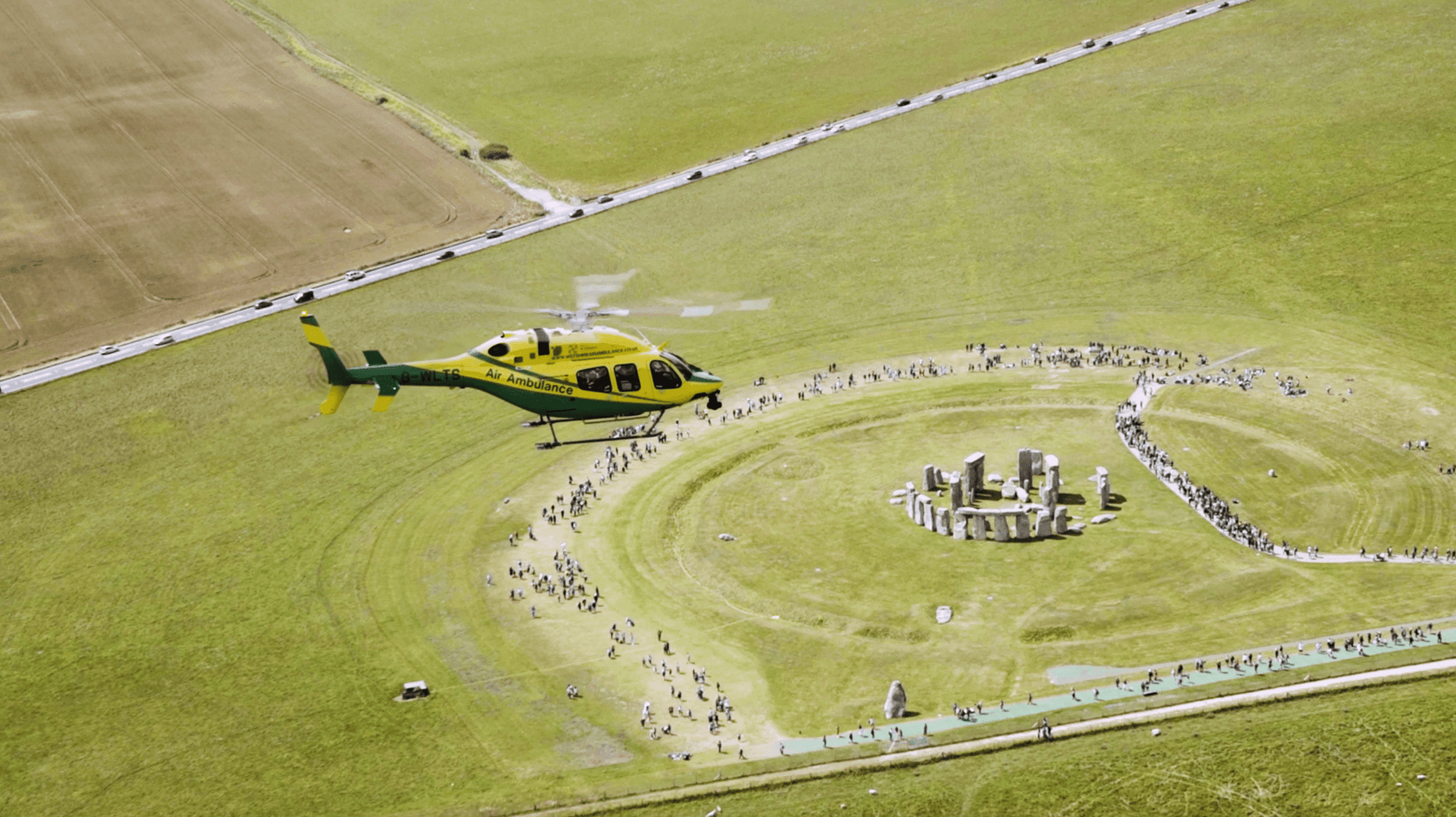 The helicopter flying over Stone Henge with lots of visitors walking around.