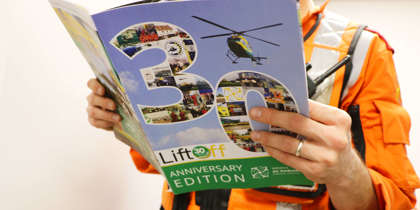 A critical care paramedic wearing an orange flight suit and reading the 30 years anniversary edition of Lift Off magazine.