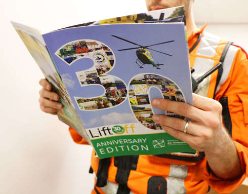 A critical care paramedic wearing an orange flight suit and reading the 30 years anniversary edition of Lift Off magazine.