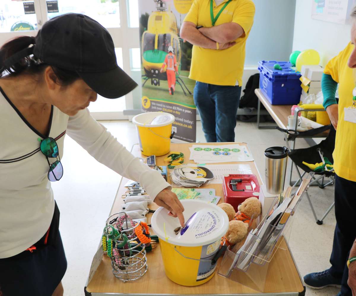 A supporter putting coins into a yellow collection bucket at a supermarket collection. There are three volunteers and table full of merchandise behind them.