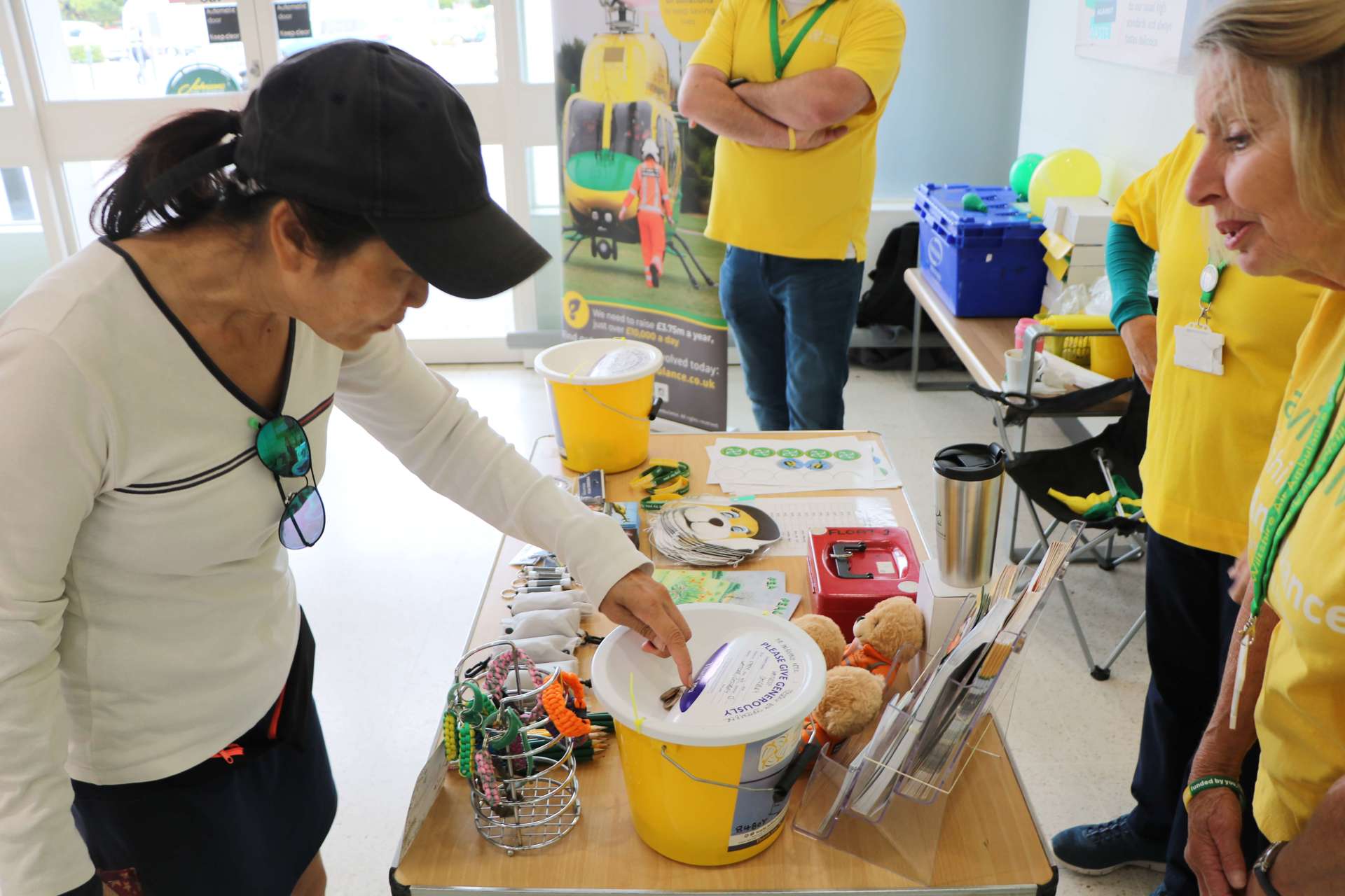 A supporter putting coins into a yellow collection bucket at a supermarket collection. There are three volunteers and table full of merchandise behind them.