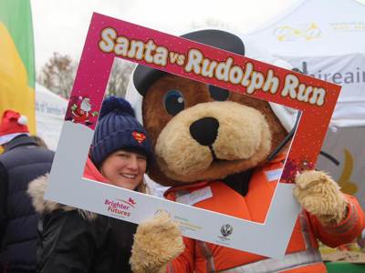 Bear mascot Wilber holding a red and white Santa vs Rudolph run selfie frame with supporter