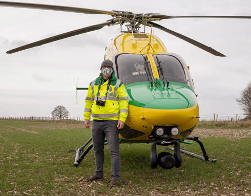 A photographer stood in front of the helicopter wearing a high vis jacket and face covering, they are also wearing a camera on a neck strap. The helicopter has landed in a green field.
