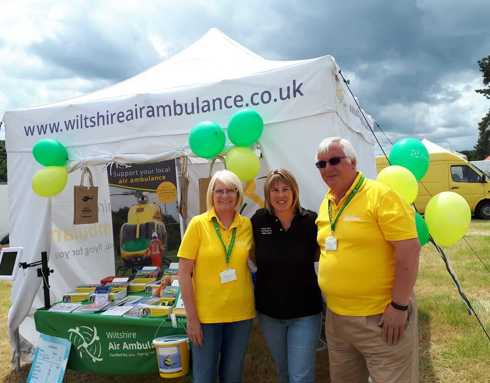 A photo of two Wiltshire Air Ambulance volunteers at an event with a Wiltshire Air Ambulance pilot. The photo is in front of a white gazebo with event merchandise.