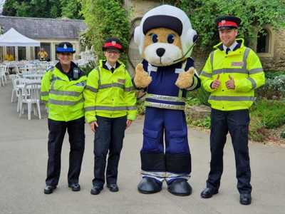 Marsha the teddy bear pilot mascot stood in a courtyard with three police officers wearing high-vis jackets.