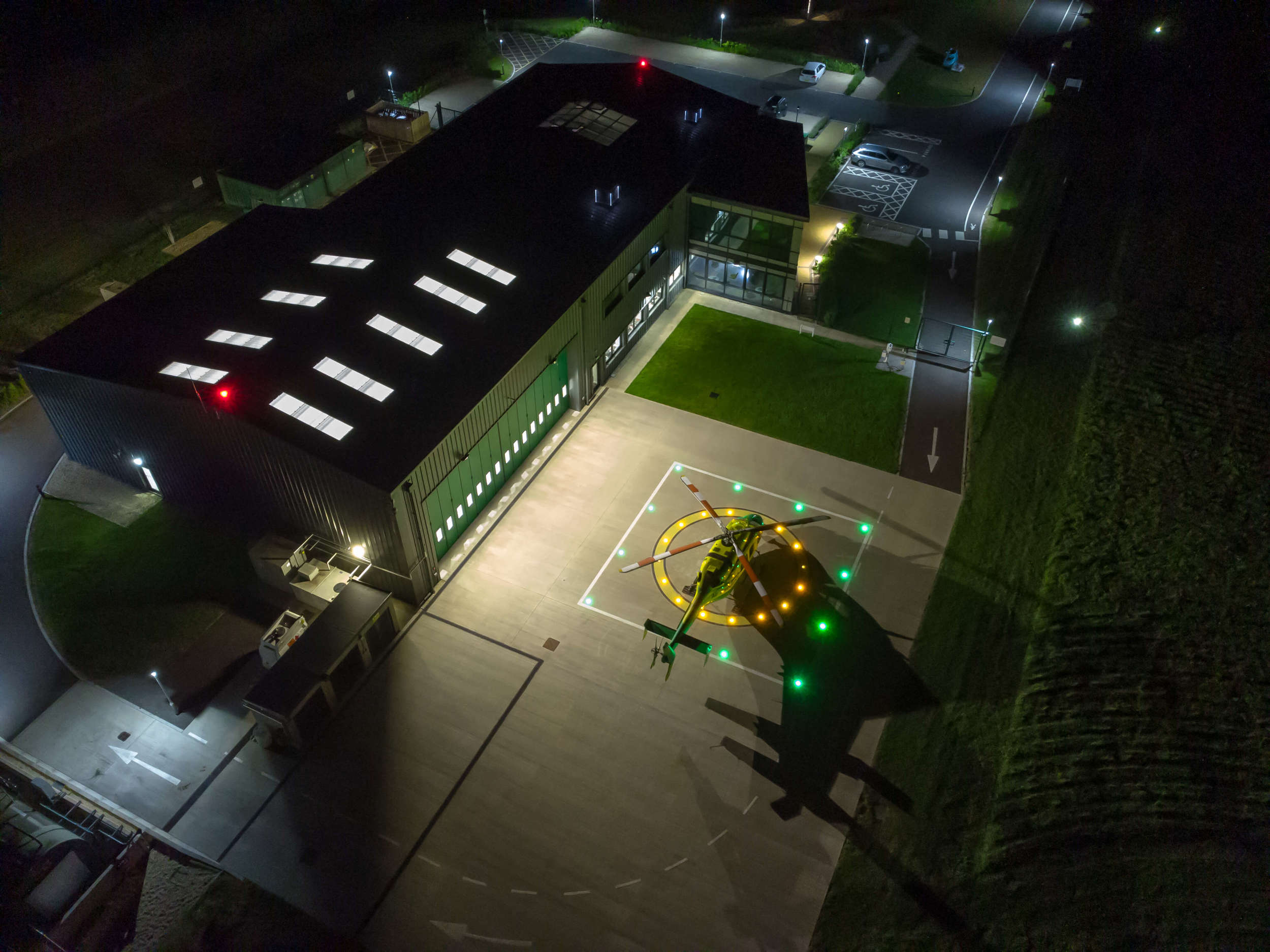 An aerial photo taken of the airbase at night. The helicopter is sat on the helipad and has bright green and yellow lights shining in a square and circle shape.