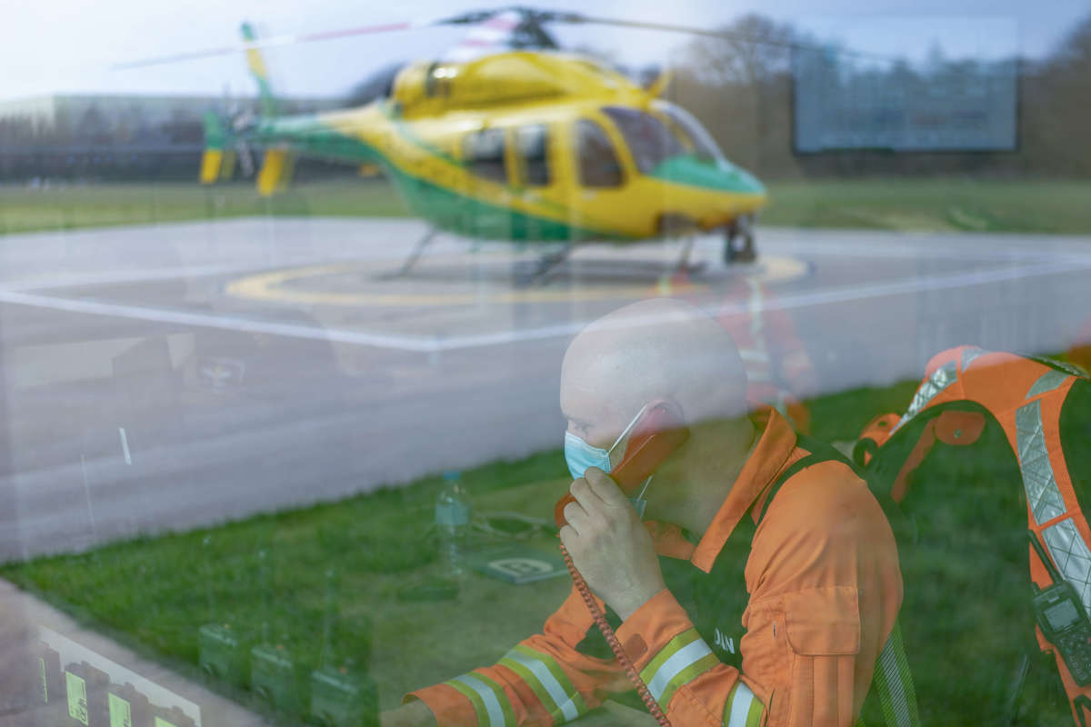 A paramedic wearing an orange flight suit answering a phone call in the flight room. There is a reflection of the helicopter on the helipad in the window.