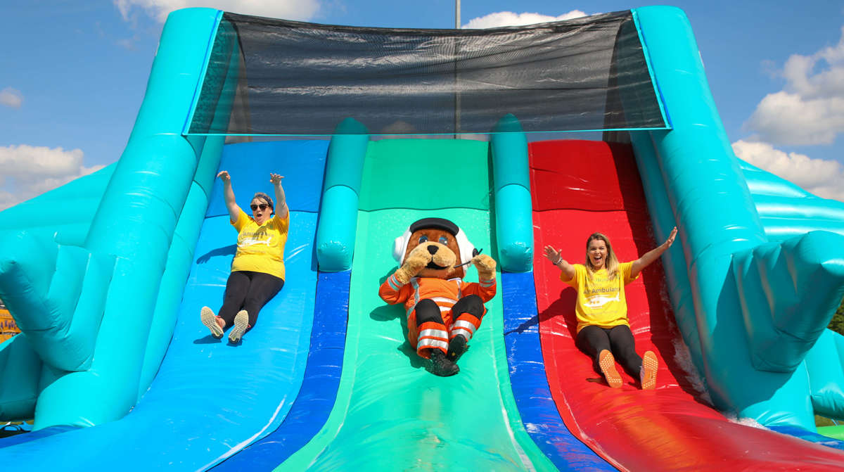 Wilber, bear mascot, sliding down an inflatable slide with two women wearing yellow Wiltshire Air Ambulance t-shirts