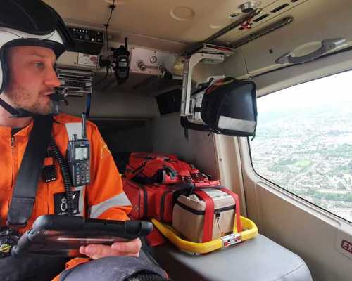 A paramedic wearing an orange flight suit answering a phone call in the flight room. There is a reflection of the helicopter on the helipad in the window.