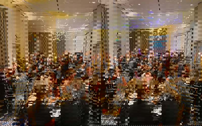 Black and gold themed charity ball. Groups of people wearing smart clothing, sitting on gold chairs around round tables