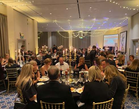 Black and gold themed charity ball. Groups of people wearing smart clothing, sitting on gold chairs around round tables