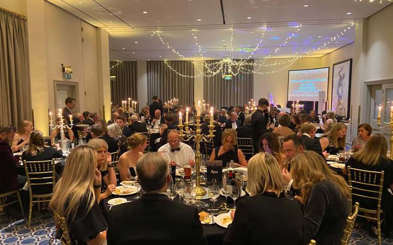 Black and gold themed charity ball. Groups of people wearing smart clothing, sitting on gold chairs around round tables