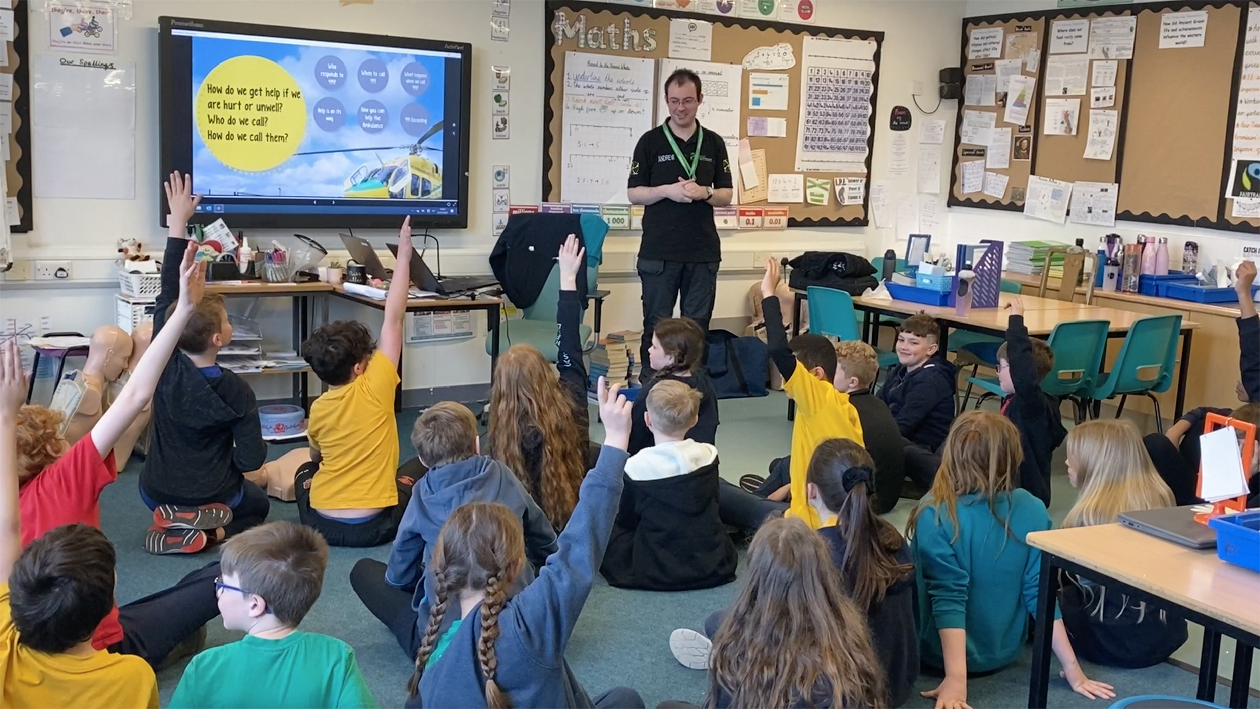 Trainer delivering emergency awareness training to a class of school children, who are sat on the floor with their hands up