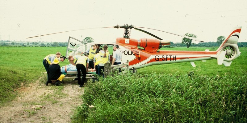 A scanned photo of a red helicopter landed in a field with police lifting a patient on a stretcher.