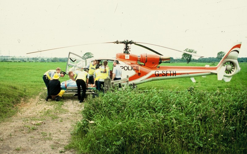 A scanned photo of a red helicopter landed in a field with police lifting a patient on a stretcher.