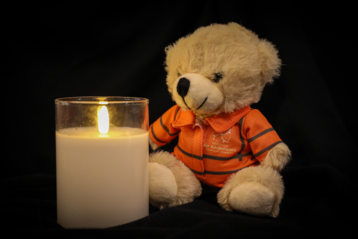 A photo of a lit candle with a small teddy bear sat next to it, wearing an orange flight suit, on a black background