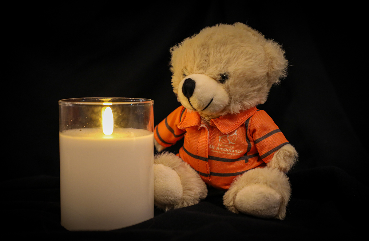 A photo of a lit candle with a small teddy bear sat next to it, wearing an orange flight suit, on a black background