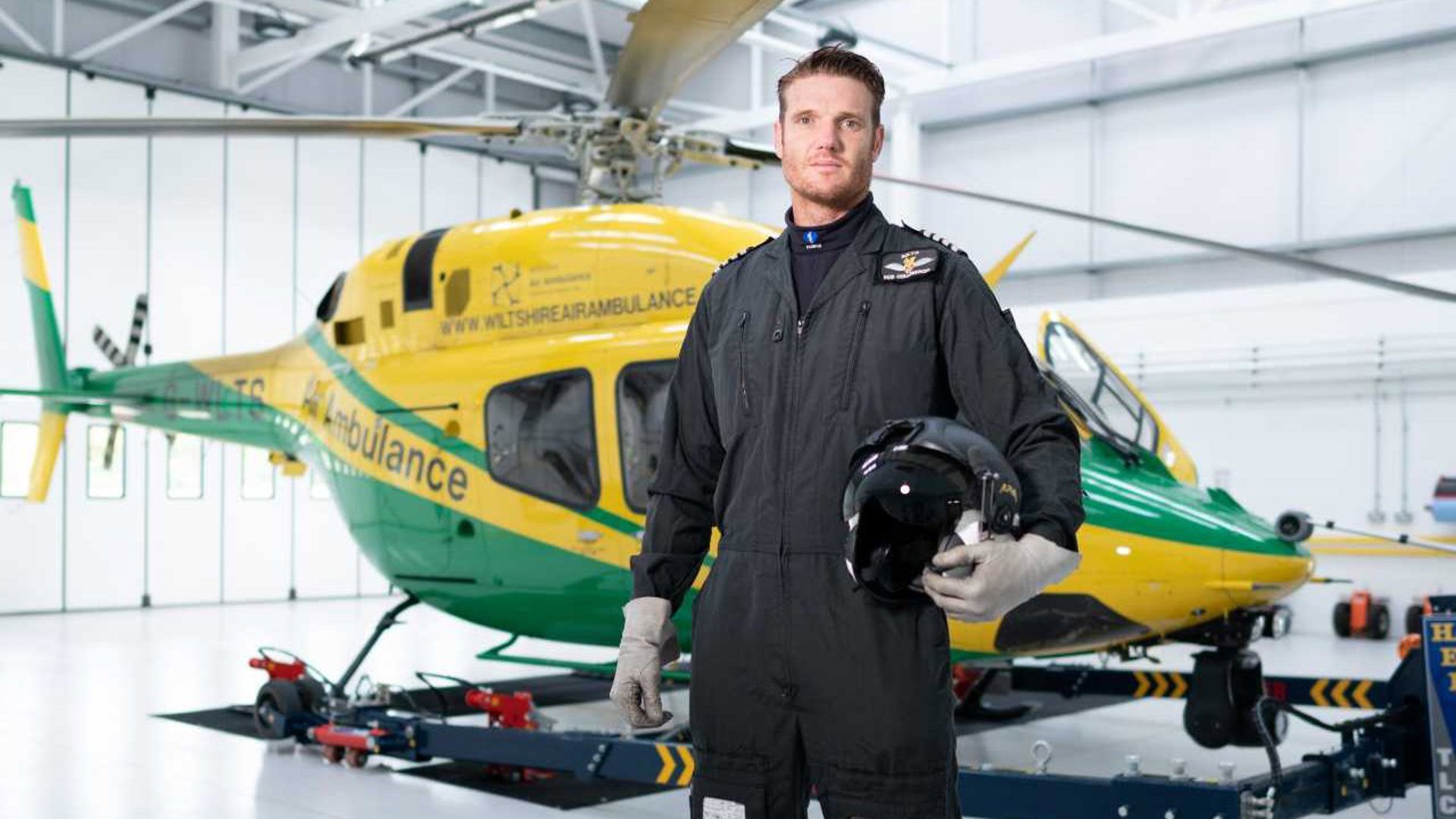 Pilot Rob Collingwood stood wearing a flight suit and holding a flight helmet in front of the helicopter in the hangar.
