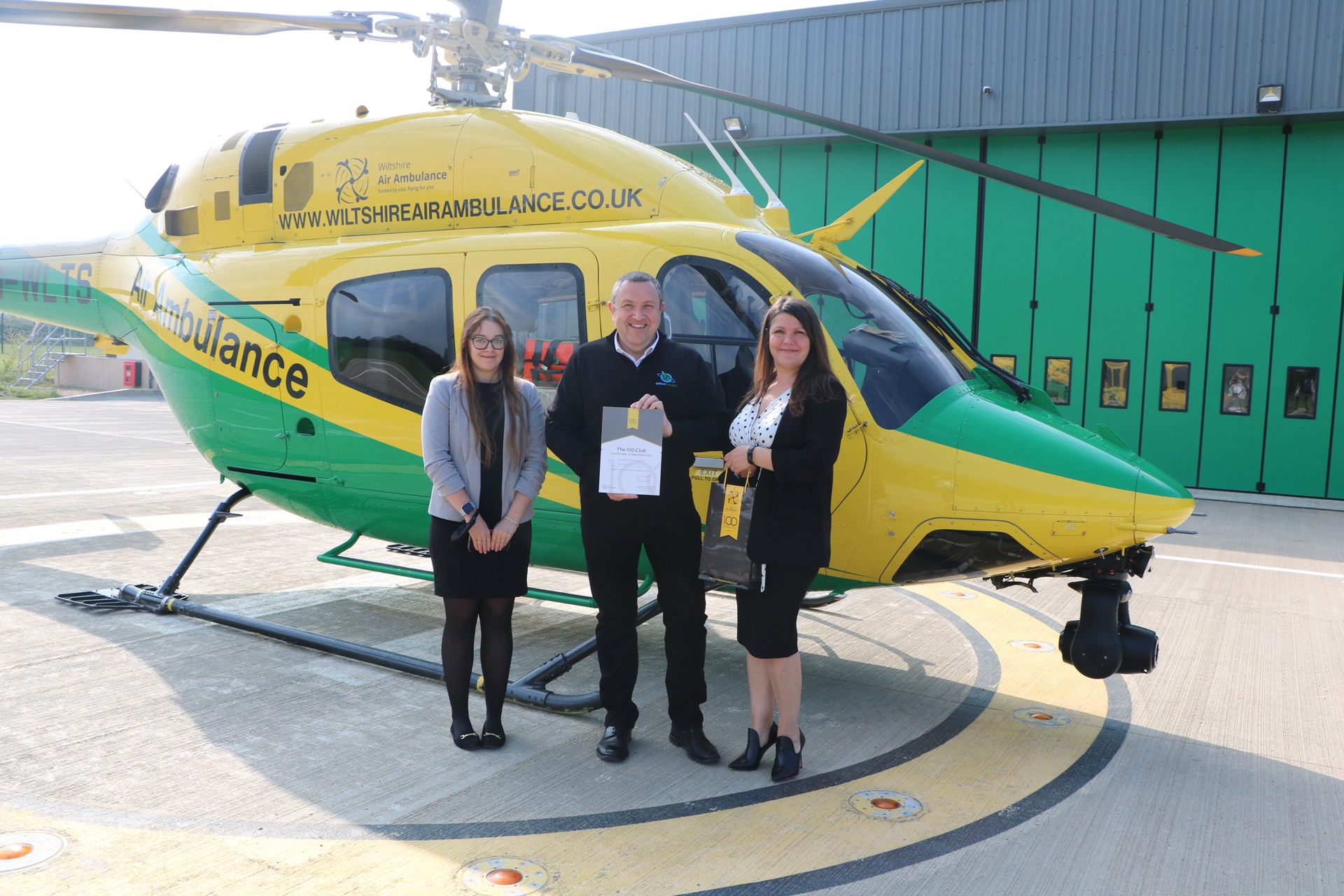 Matt Goodall and Office Evolution colleagues holding 100 club branded certificate and gift bag, standing in front of the helicopter