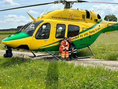 A photo of a paramedic and pilot smiling at the camera whilst perching on the step of the Wiltshire Air Ambulance's yellow and green helicopter