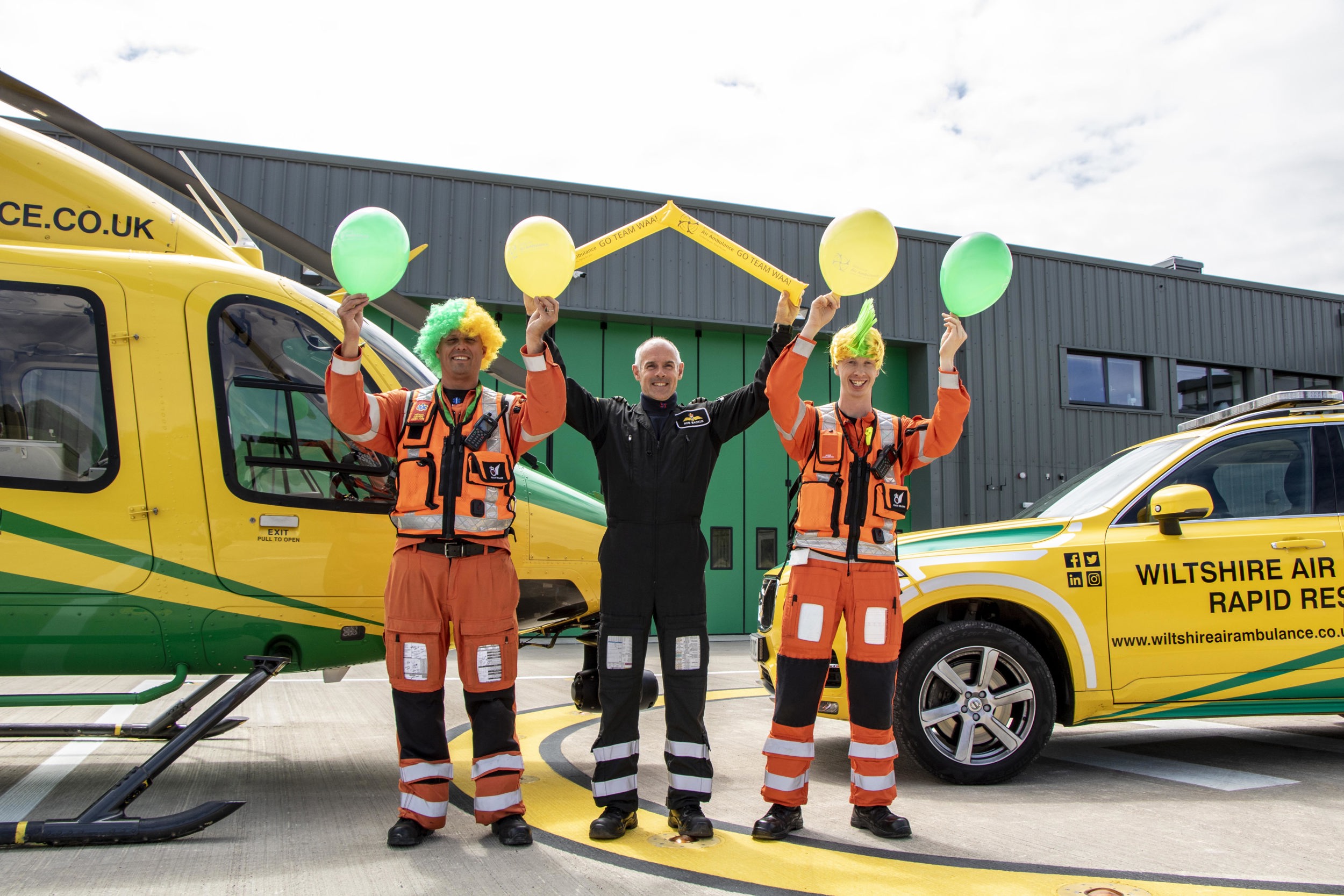 Paramedic Rich Miller wearing a yellow and green curly wig and holding yellow and green balloons, Rob Backus holding yellow Team WAA cheer sticks and Craig Wilkins wearing a yellow and green mohawk wig and holding yellow and green balloons