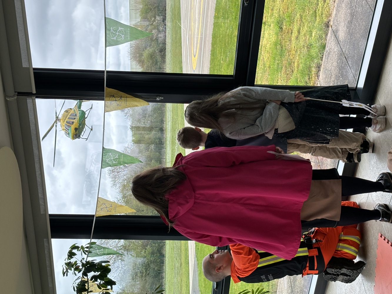An adult and two children watching a yellow and green helicopter take off, with a critical care paramedic wearing orange
