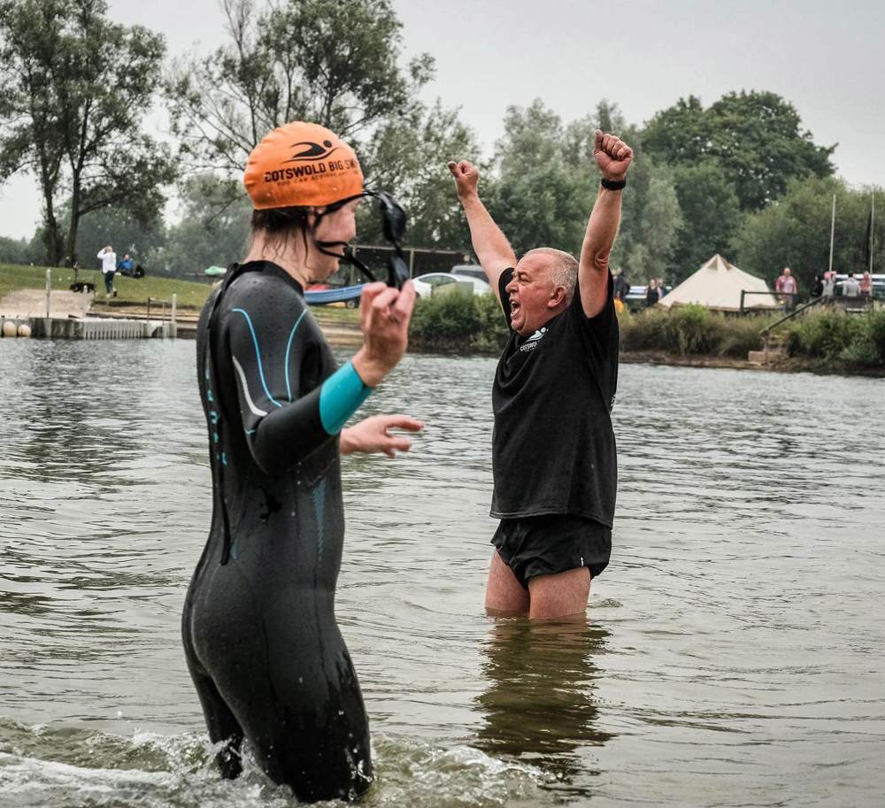 A swimmer wearing a wet suit and orange swim hat, standing knee deep in water, with a man behind wearing a t-shirt and shorts with his arms in the air cheering people on